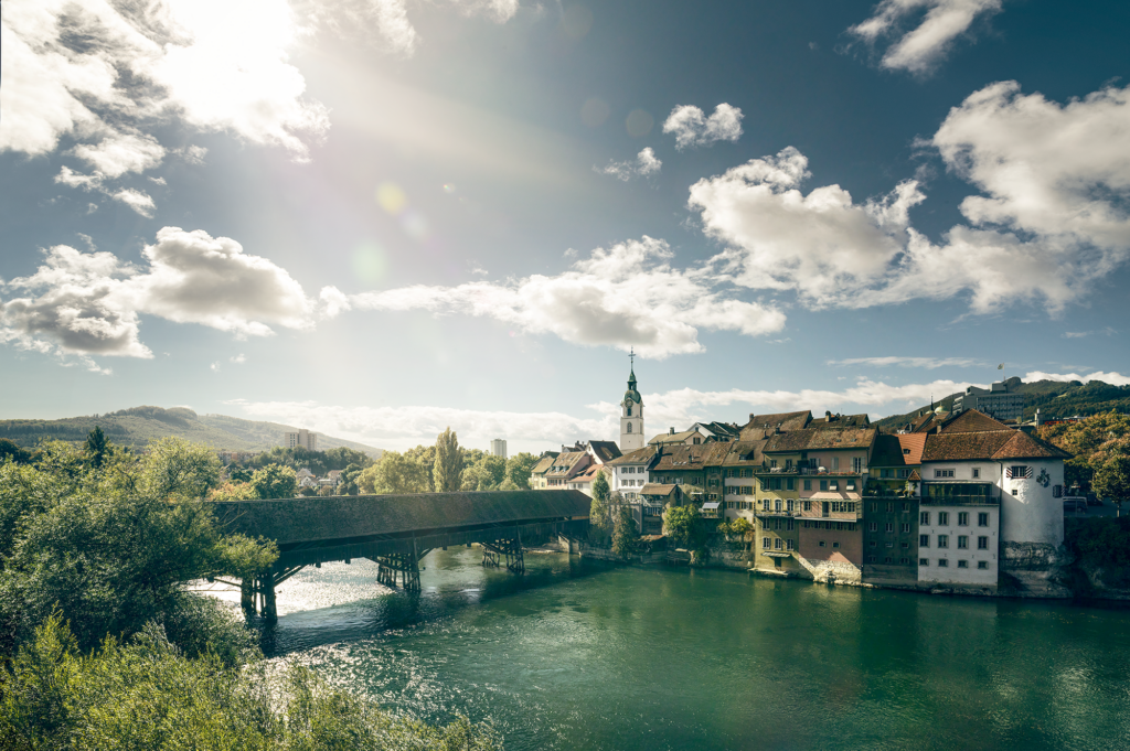 Fluss mit Brücke und Stadt im Hintergrund, Himmel mit Wolken und Sonne.