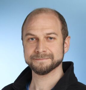 Mann mit Bart, blauen Augen, schwarzem Pullover vor blauem Hintergrund.