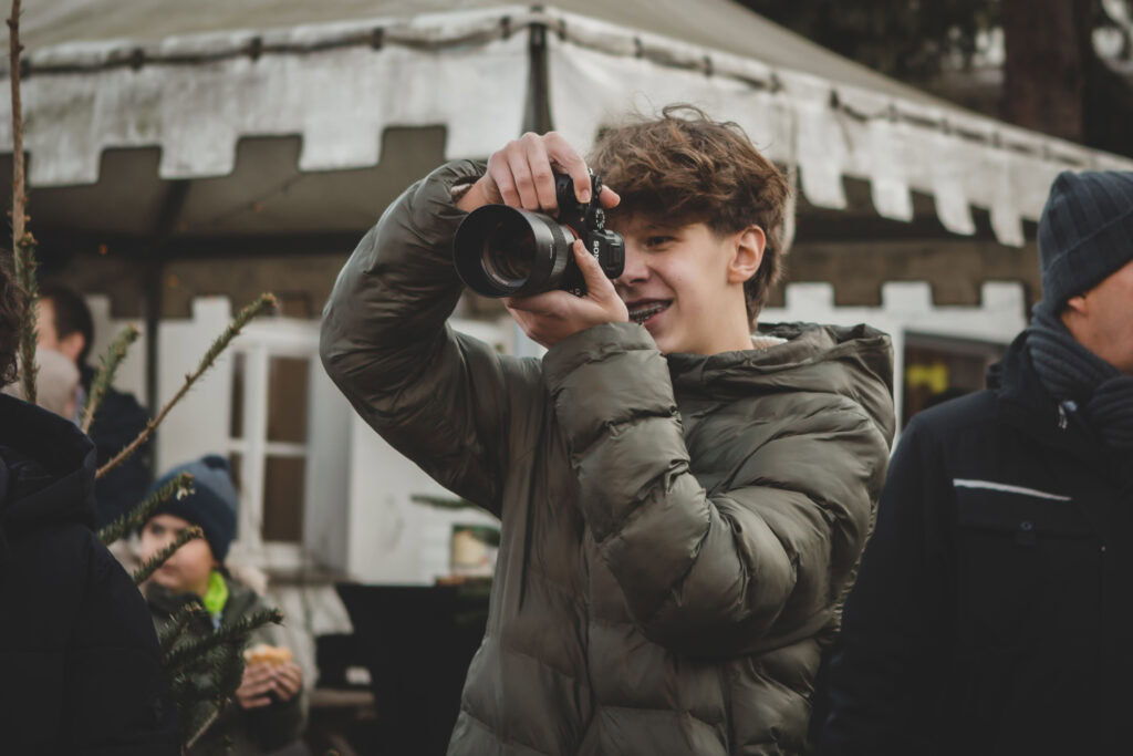 Junger Mann mit Brille hält Kamera und macht Fotos bei winterlichem Weihnachtsmarkt in Essen, umgeben von festlich geschmückten Verkaufsständen und Freunden, ideal für christliche Veranstaltungen und Gemeindeleben.