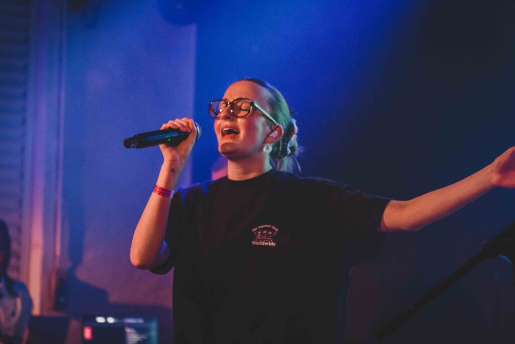 Eine junge Frau mit Brille singt in ein Mikrofon, mit ausgestrecktem Arm.