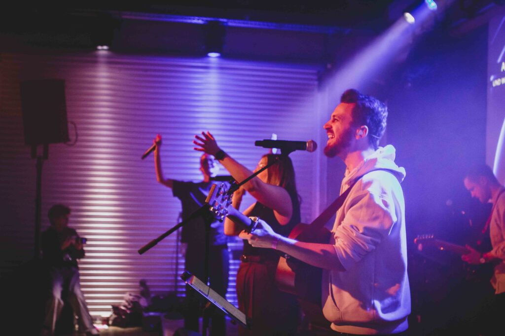 Musiker singt und spielt Gitarre auf der Bühne, mit anderen Musikern im Hintergrund.