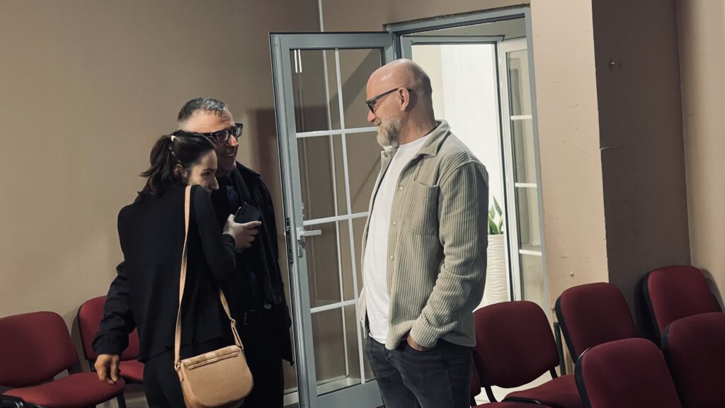 Three people are standing and talking in a room with red chairs and a glass door.