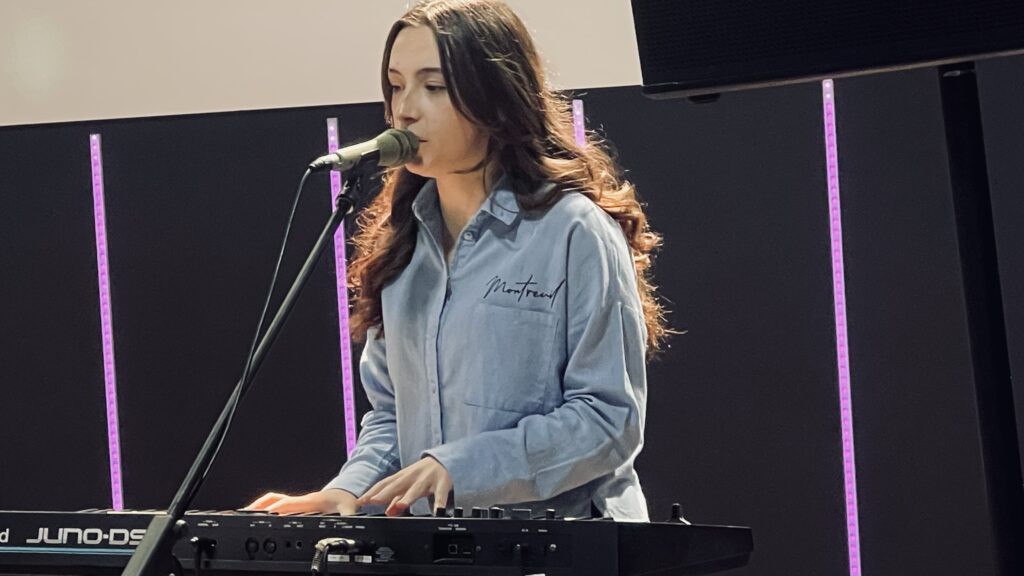 A woman with long dark hair singing into a microphone while playing a keyboard.