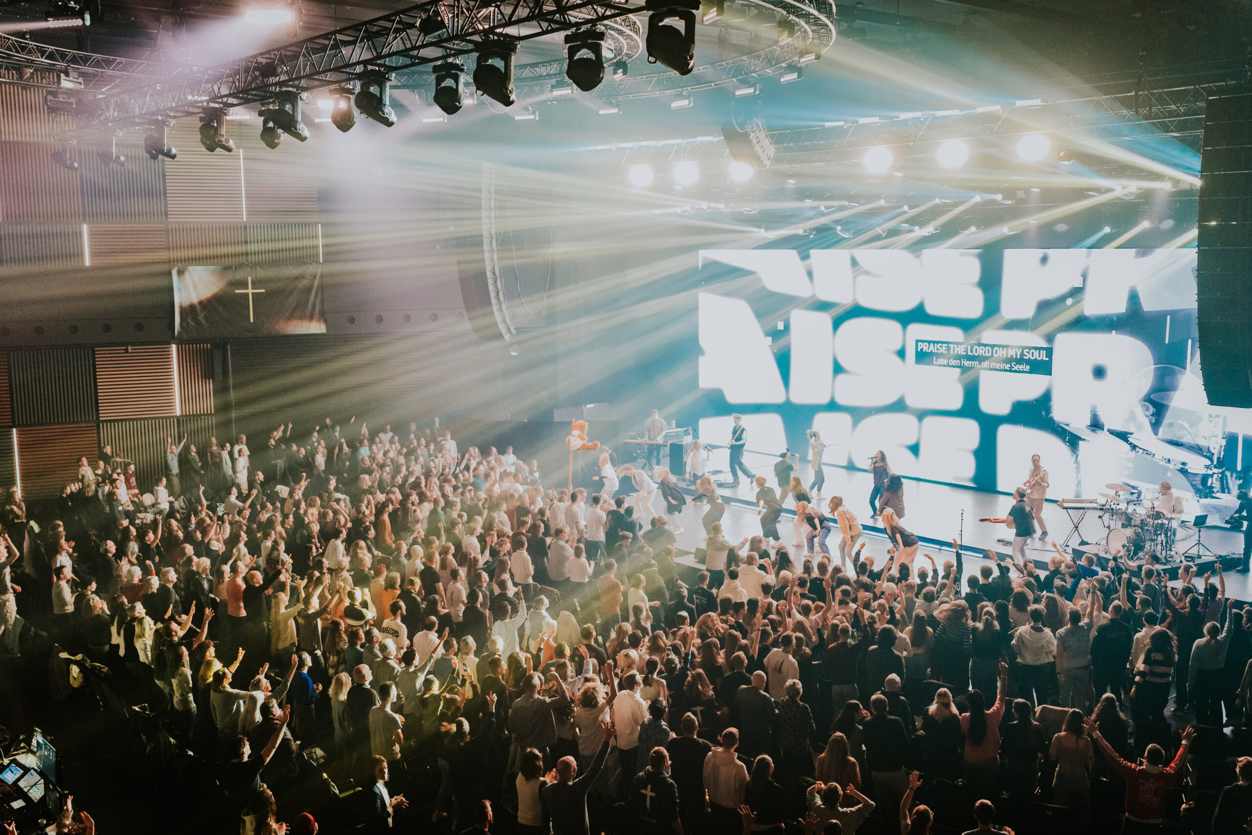 A large crowd gathered in front of a stage with bright lights and a large screen displaying "RISE NA.