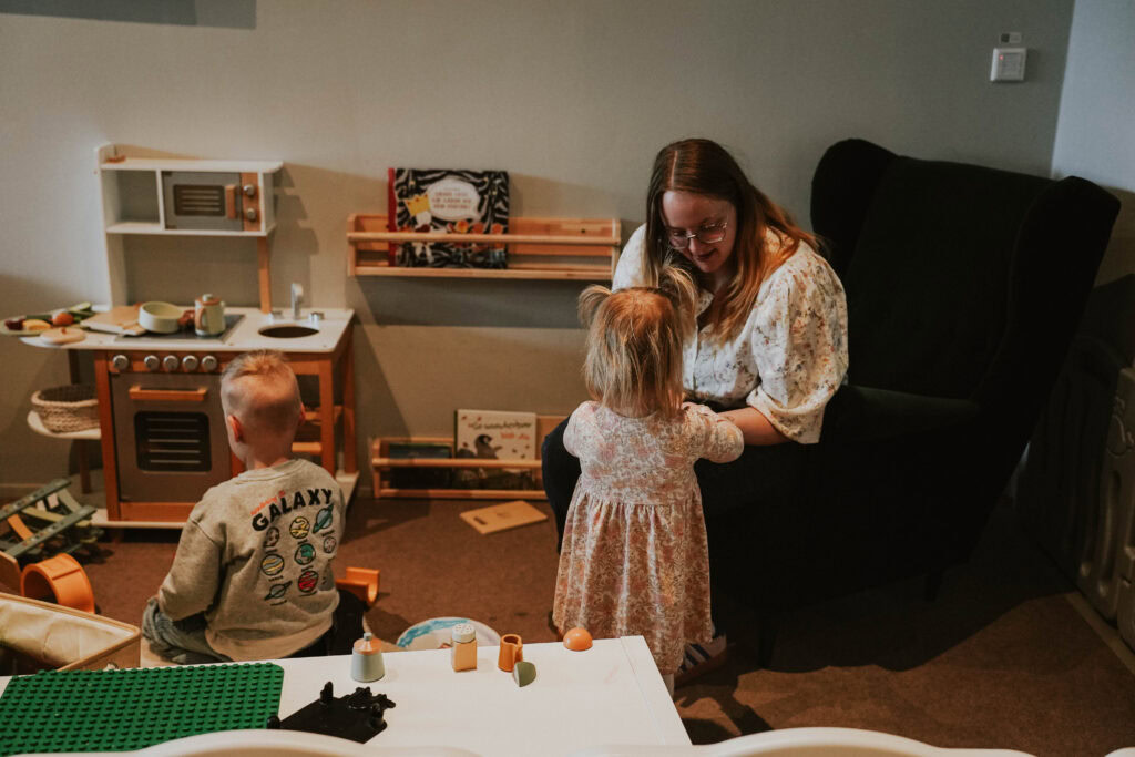Kinder spielen im Spielzimmer, eine Frau sitzt auf einem schwarzen Sessel und umarmt ein kleines Mäd.