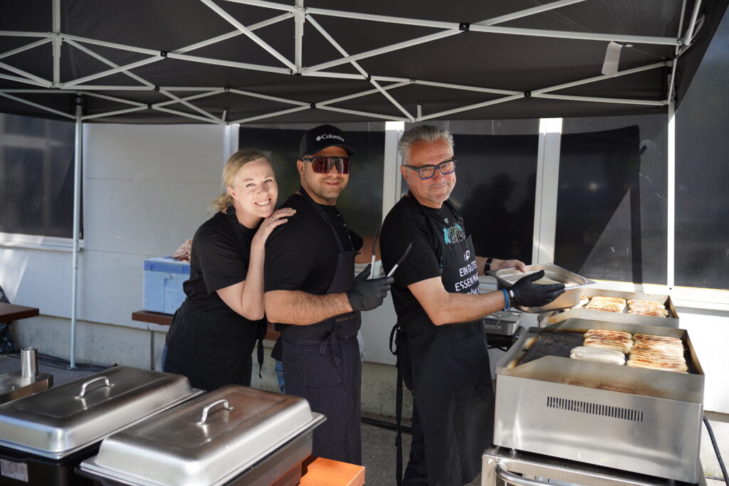Drei Personen grillen Essen an einem Außenkocher unter einem Zelt.