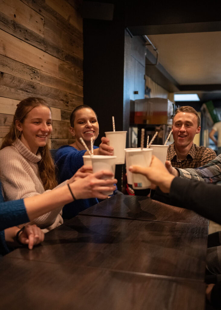 Einladende Gruppe junger Menschen, die gemeinsam Getränke in einem modernen Café oder Restaurant genießen, mit freundlicher Atmosphäre und Holzdekoration im Hintergrund.