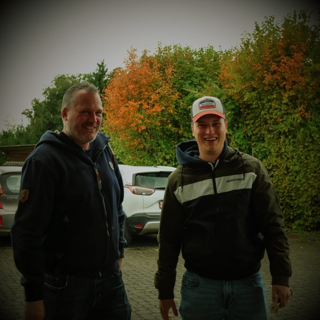 Der Mann und die junge Frau lachen gemeinsam im Freien vor einem Parkplatz mit herbstlicher Laubfärbung.