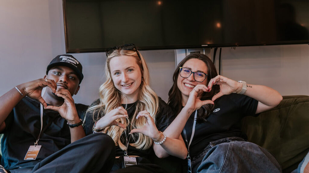 Three individuals sitting on a couch, making heart shaapes with their hands.