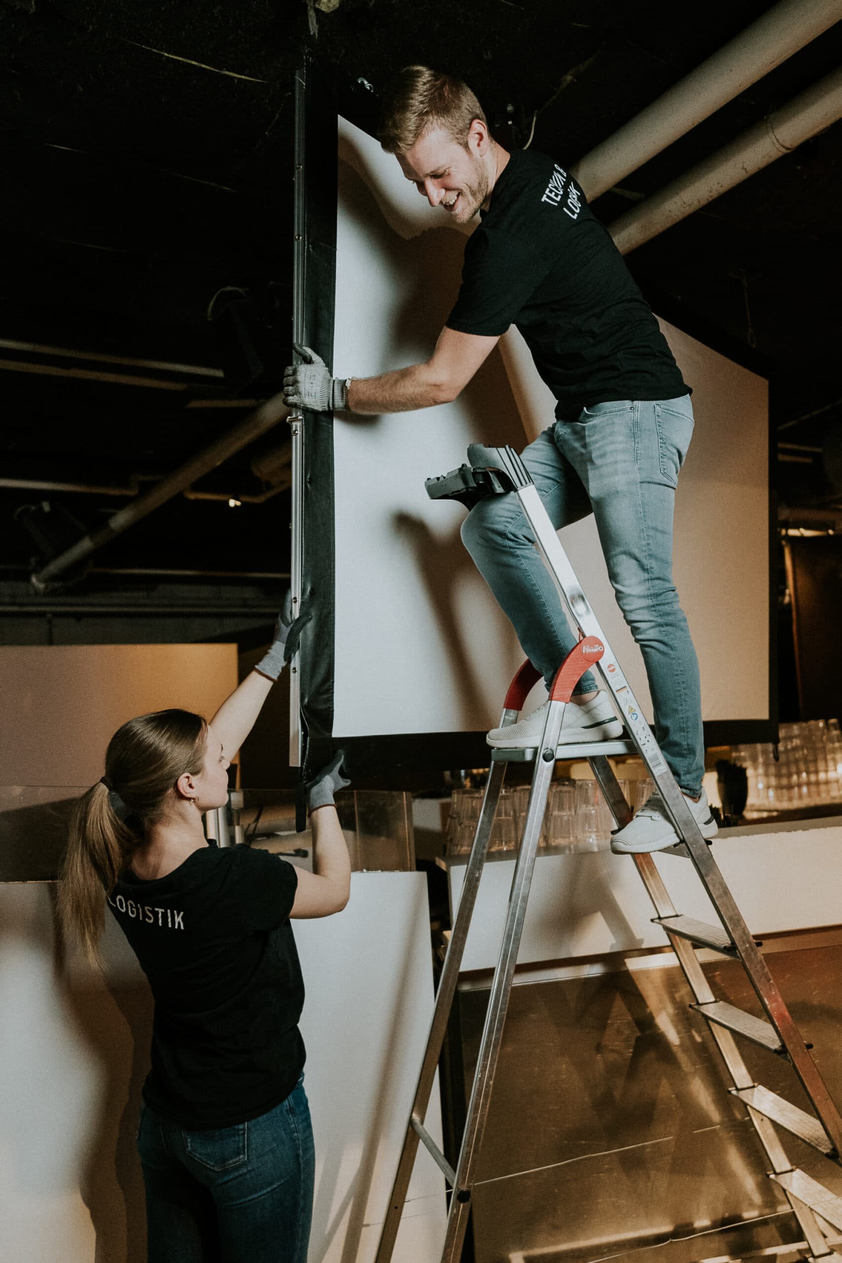 Montage eines Displays oder Filmsystems mit zwei Technikern in einem Veranstaltungsraum. Ein Mann steht auf einer Leiter und befestigt das System, eine Frau unterstützt ihn. Professionelle Veranstaltungstechnik, Eventplanung, audiovisuelle Technik, Eventmanagement.