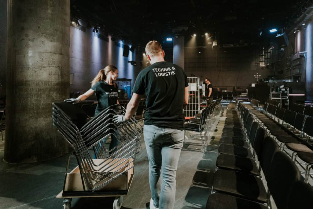 Innenraum eines Kirchen- oder Veranstaltungsraums, Team von Technikern mit T-Shirts "Technik & Logistik" beim Aufbau, Monitore und Stühle, Vorbereitungen für einen Gottesdienst oder Event, professionelle Veranstaltungstechnik.