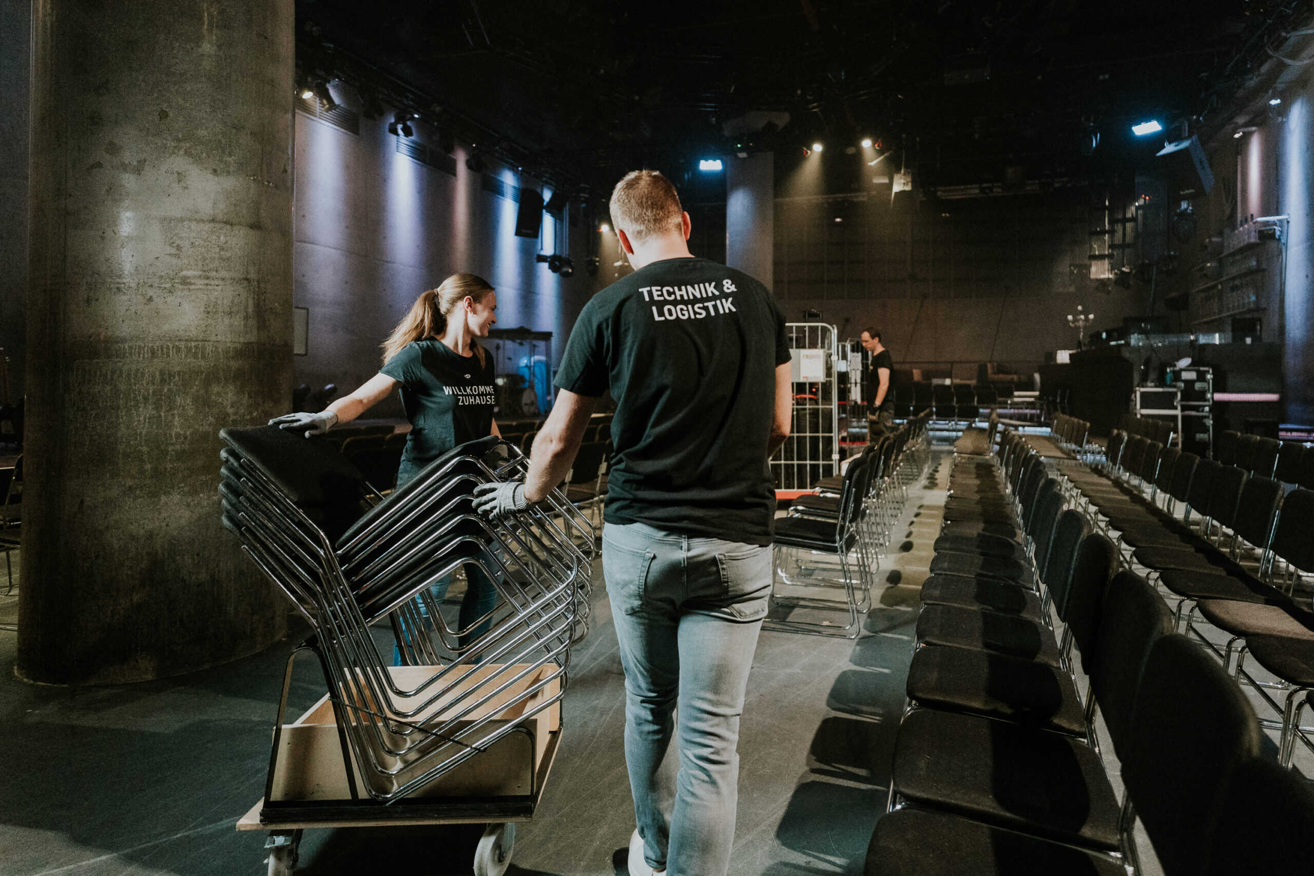 Innenraum eines Kirchen- oder Veranstaltungsraums, Team von Technikern mit T-Shirts "Technik & Logistik" beim Aufbau, Monitore und Stühle, Vorbereitungen für einen Gottesdienst oder Event, professionelle Veranstaltungstechnik.