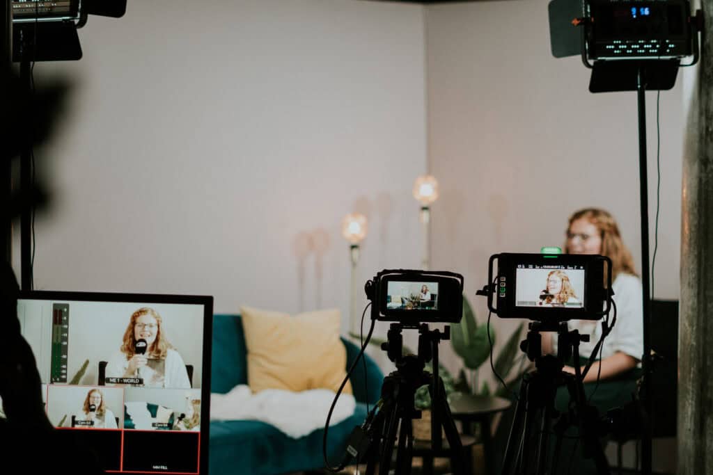 Ehefrau im Videochat während einer Live-Übertragung, auf einer Couch mit Kissen, in Hintergrund Pflanzen und Stehlampen, Kamera- und Lichttechnik im Einsatz, professionelle Aufnahme für Online-Gottesdienst oder Event.