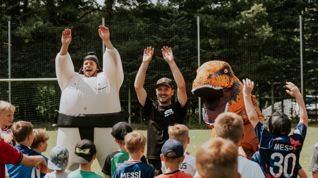 Kinder und Erwachsene feiern auf einer Sportanlage, einige in Kostümen, mit erhobenen Armen.