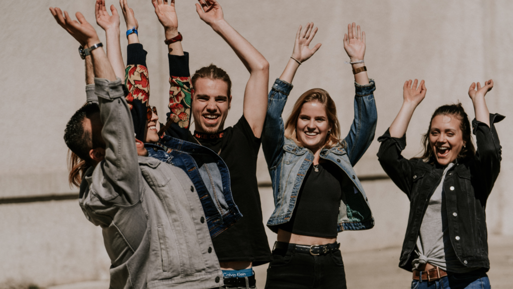 Fünf junge Menschen stehen im Freien, lachen und heben die Arme in die Luft.