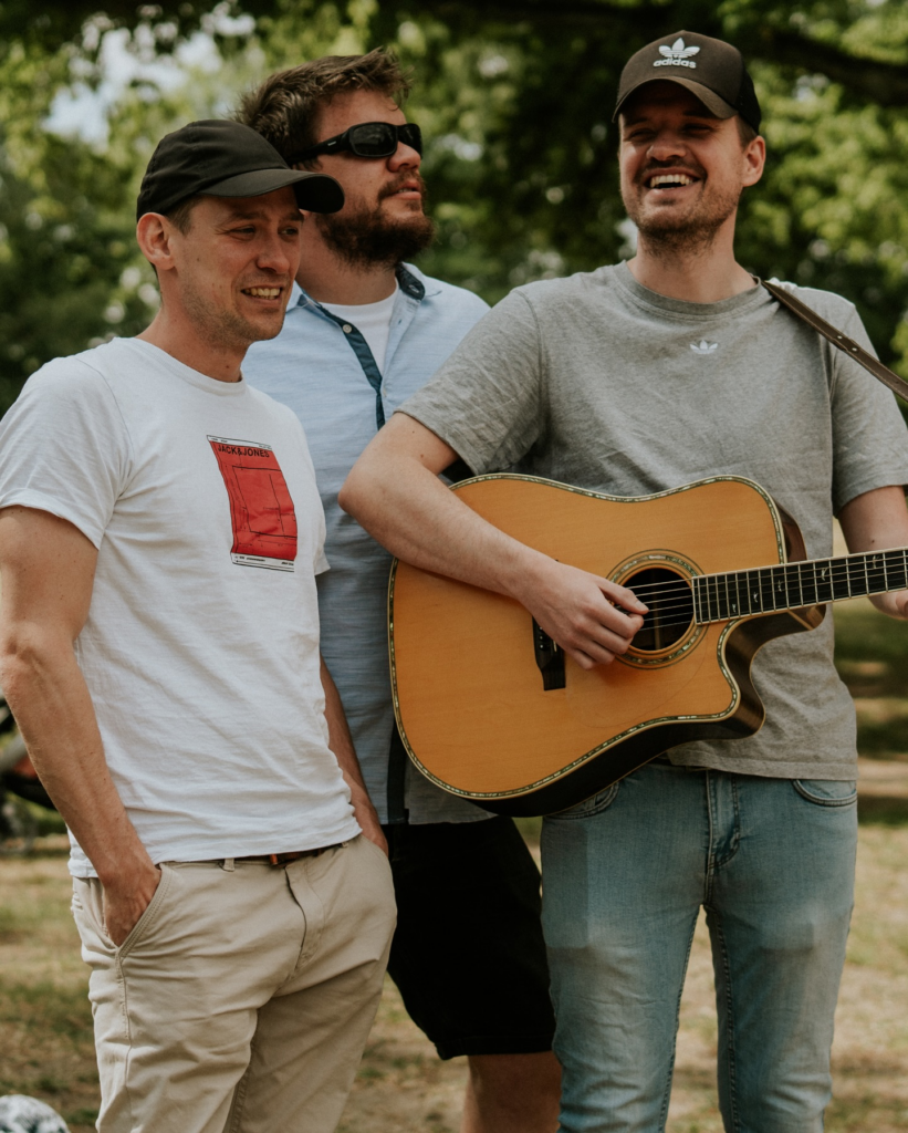 Drei Männer im Park, einer spielt Gitarre, alle lachen.