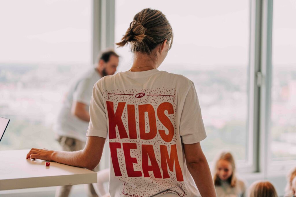Mädchen mit "Kids Team" T-Shirt bei ICF Hamburg, engagiert in Kinderbetreuung und Gemeinschaftsaktivitäten, in hellem Raum mit großen Fenstern und anderen Kindern im Hintergrund.