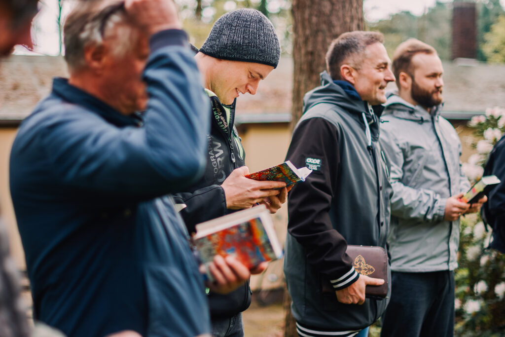 Gruppe von Menschen beim Gemeindetreffen im Freien, die Bibeln und Bücher halten, in einer natürlichen Umgebung mit Bäumen im Hintergrund.