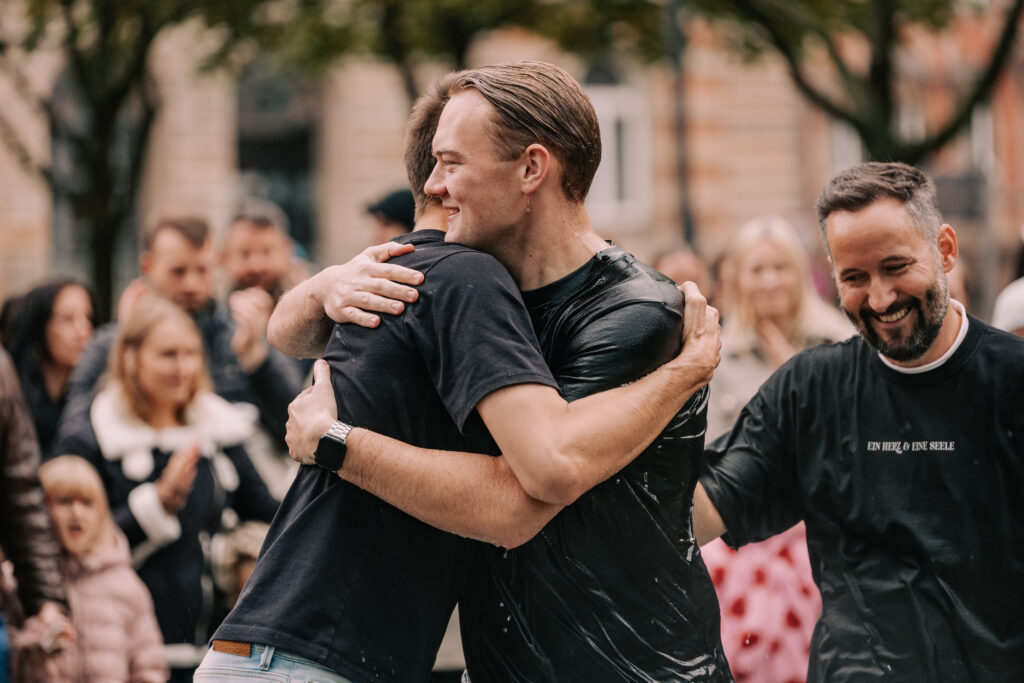 Zwei Männer umarmen sich herzlich bei einem christlichen Gemeinschafts-Event in Hamburg, umgeben von einer Gruppe lachender Menschen im Freien.