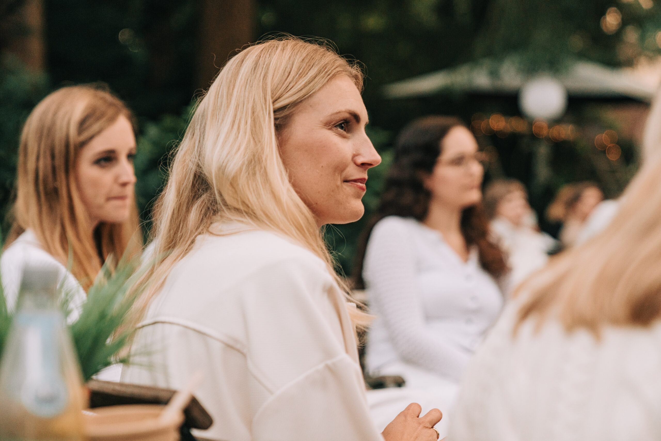 Drei Frauen sitzen im Freien bei einer Veranstaltung, eine Frau im Vordergrund blickt nach rechts.