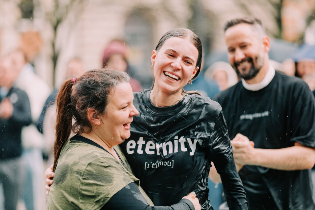 Braut in schwarzem Kleid mit "Eternity" auf der Brust, umarmt eine Frau, im Hintergrund weitere Gäst.