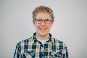 Ein Mann mit Brille, blondem, lockigem Haar und einem karierten Hemd lächelt.