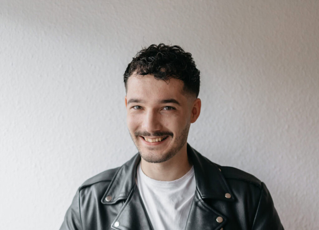 Junger Mann in schwarzer Lederjacke, lächelnd, vor weißer Wand, modebewusst, urbane Atmosphäre, Lifestyle, junge Menschen, modischer Stil.