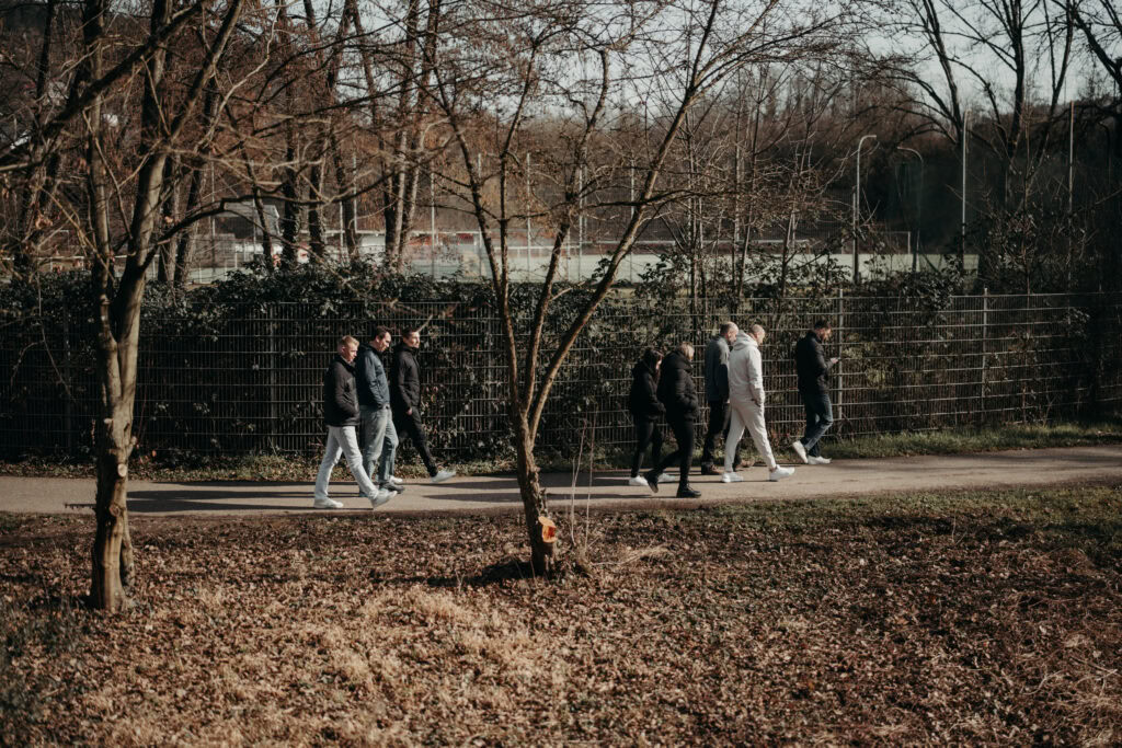 Sieben Menschen gehen auf einem Weg im Park, Bäume ohne Blätter im Hintergrund.