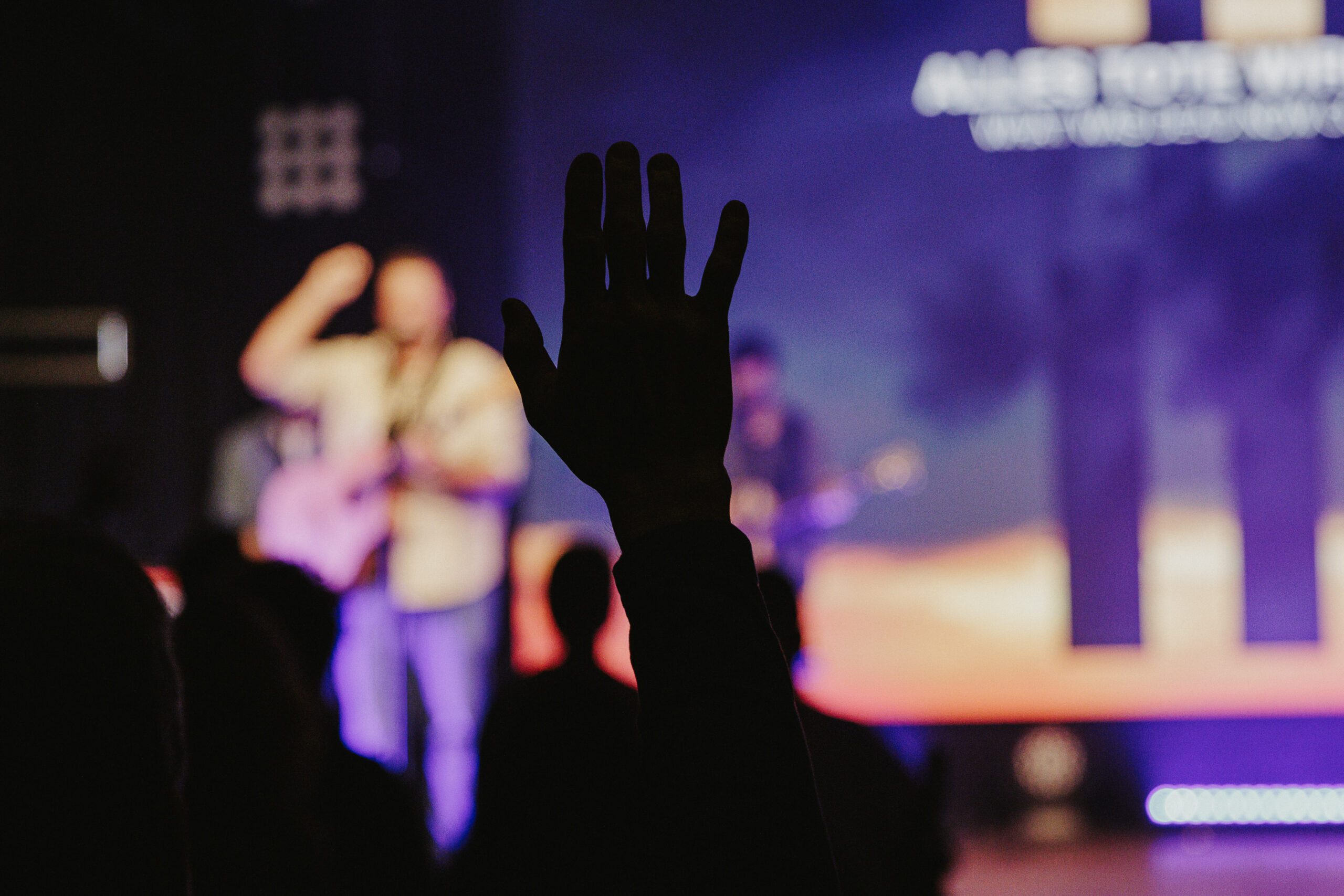 Menschen beteiligen sich an einer christlichen Glaubensveranstaltung, Fokus auf eine erhobene Hand in der Dunkelheit, mit einer Bühne und Musik im Hintergrund, Spirituelle Gemeinschaft und Worship im Mittelpunkt.