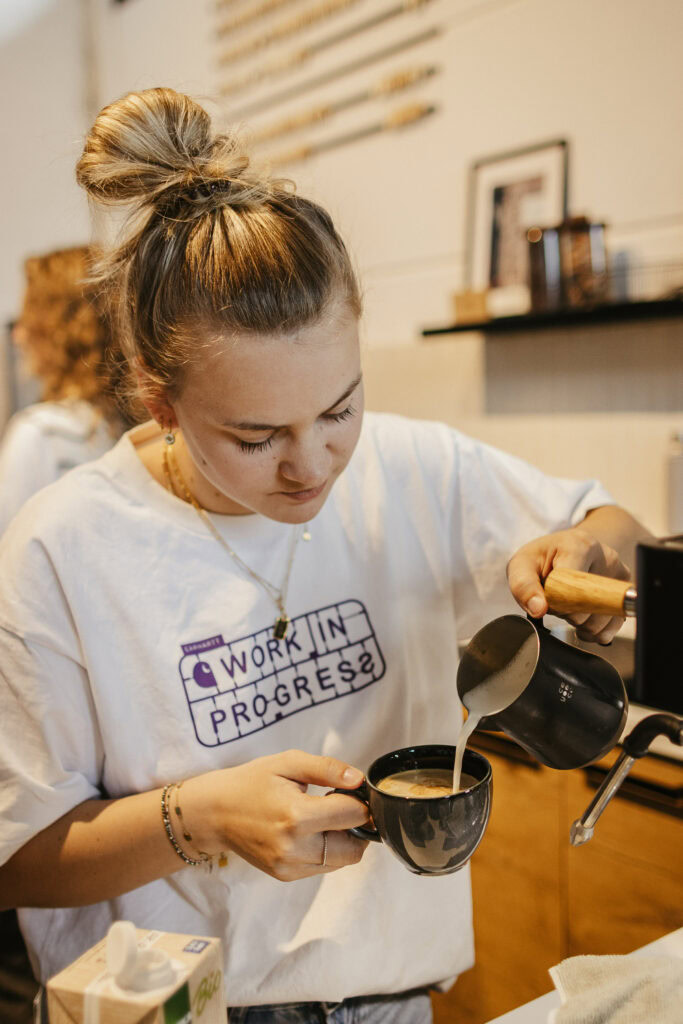 Alt-Text: Junge Frau bereitet Kaffee zu, gießt milch in eine Tasse, trägt ein weißes T-Shirt mit "Work in Progress" Schriftzug, typische gemütliche Innenraum-Atmosphäre.