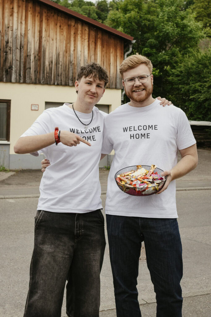 Zwei junge Männer tragen T-Shirts mit "Welcome Home" und teilen Süßigkeiten, feiern Gemeinschaft und Freundschaft bei einer christlichen Veranstaltung in Herrenberg.