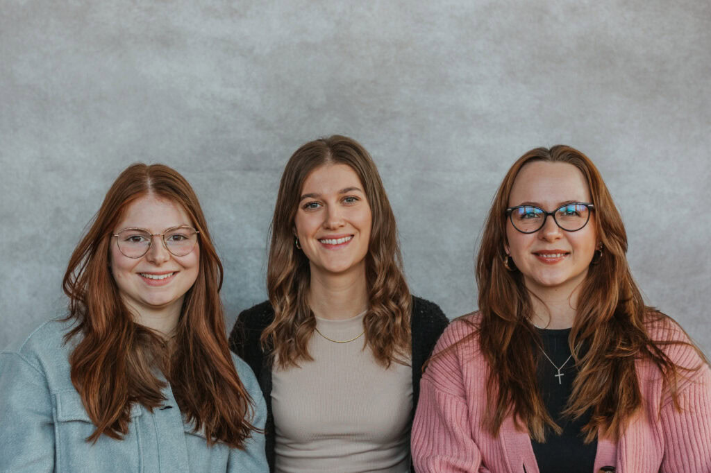 Alt: Drei lachende Frauen mit Brillen und moderner Kleidung vor einer grauen Wand, symbolisierend Gemeinschaft und Vielfalt bei ICF XY in Herrenberg.