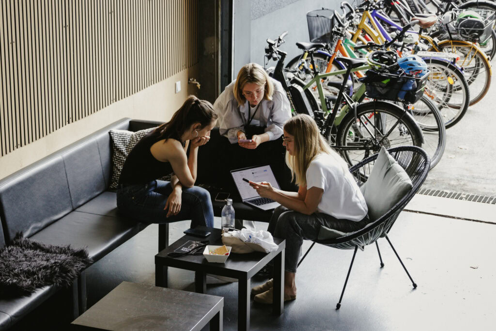 Drei Frauen sitzen im Café und diskutieren gemeinsam, während sie auf einem Tablet schauen, mit Fahrrädern im Hintergrund.