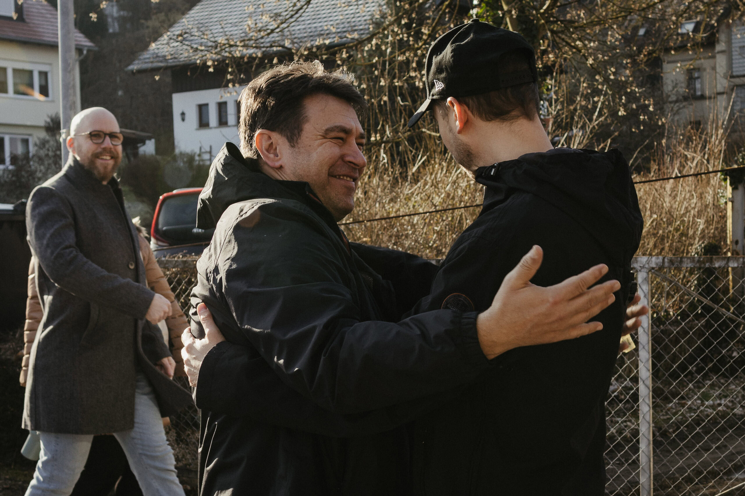 Zwei Männer begrüßen sich herzlich im Freien, während im Hintergrund ein weiterer Mann lächelnd zuschaut. Die Szene zeigt Gemeinschaft und Freude bei einem Treffen im Freien.