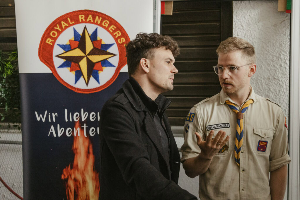 Zwei Männer sprechen vor einem Royal Rangers-Logo, einer trägt ein Scout-Kleid.