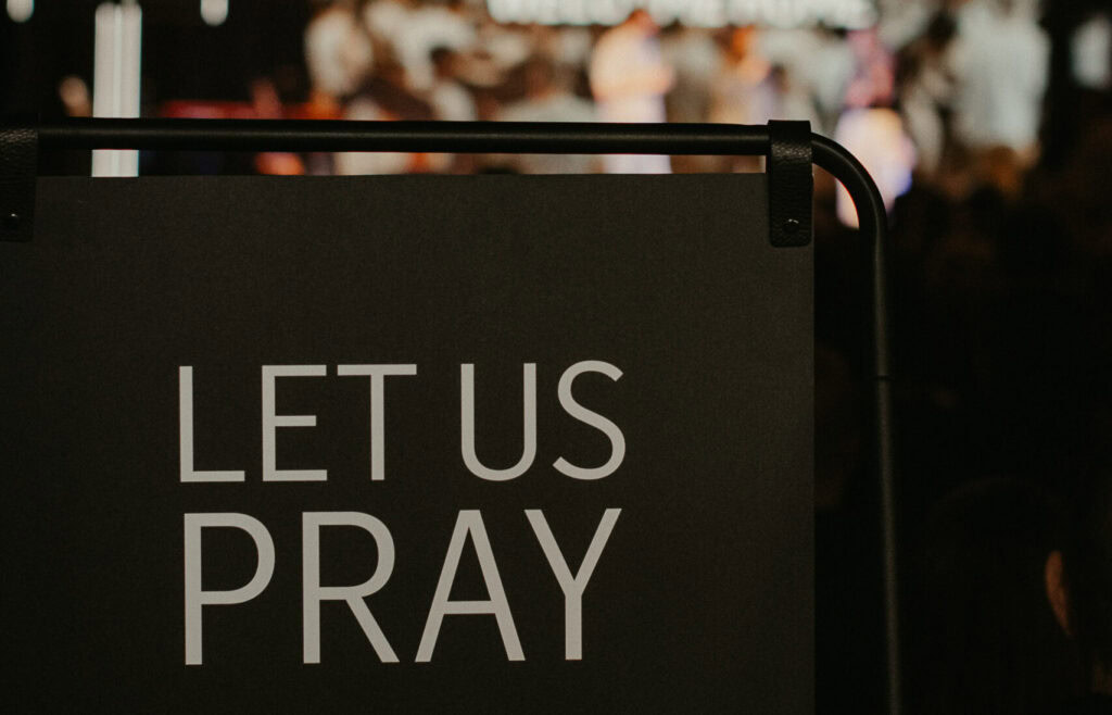 Schwarzer Stehposter mit weißen Text "LET US PRAY" und Gebetshand-Symbol.