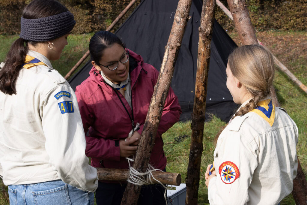 Drei Frauen in Scout-Uniformen stehen vor einem aufgebauten Zelt aus Holzstämmen.