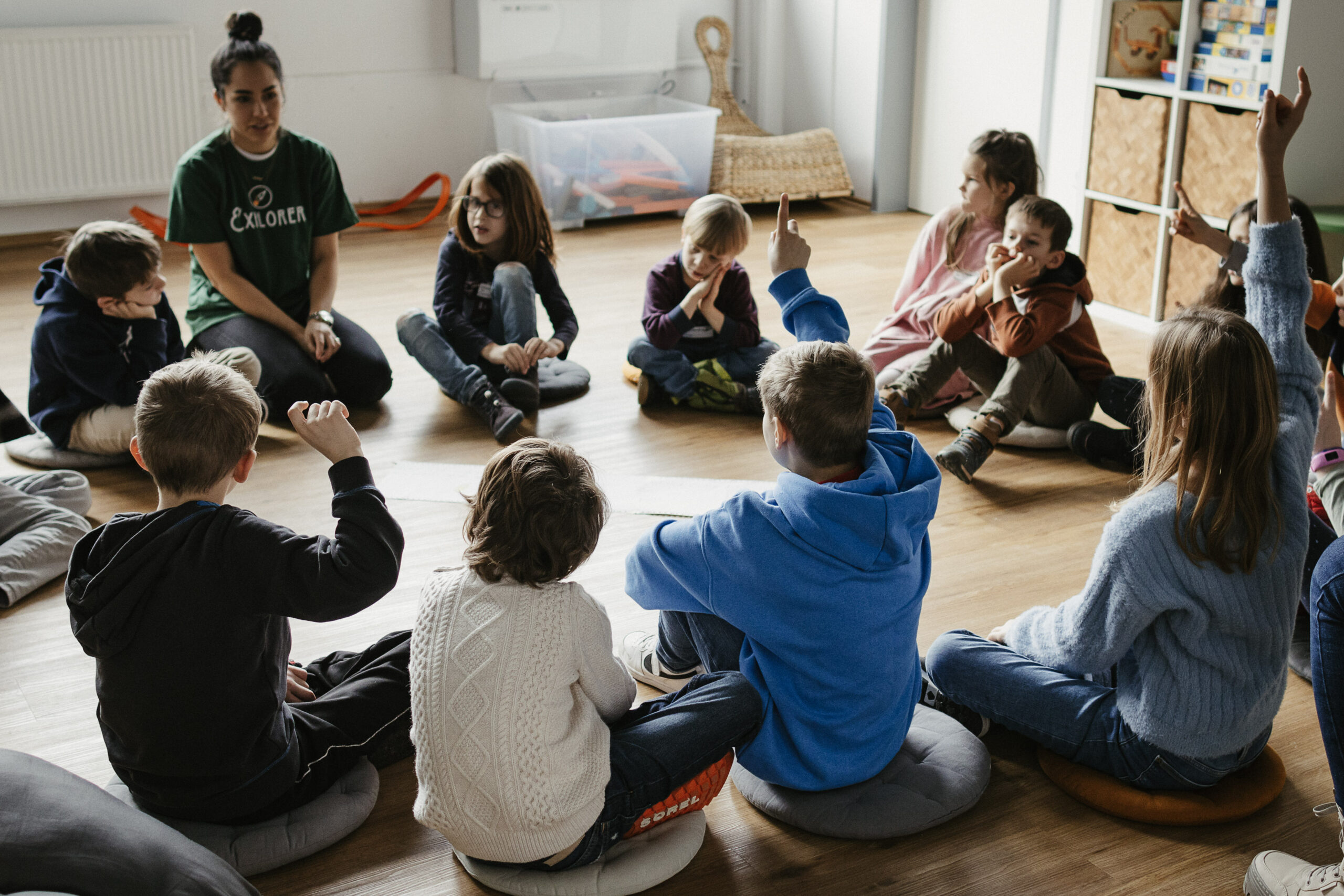 Kinder sitzen im Kreis auf dem Boden, einige heben die Hand.