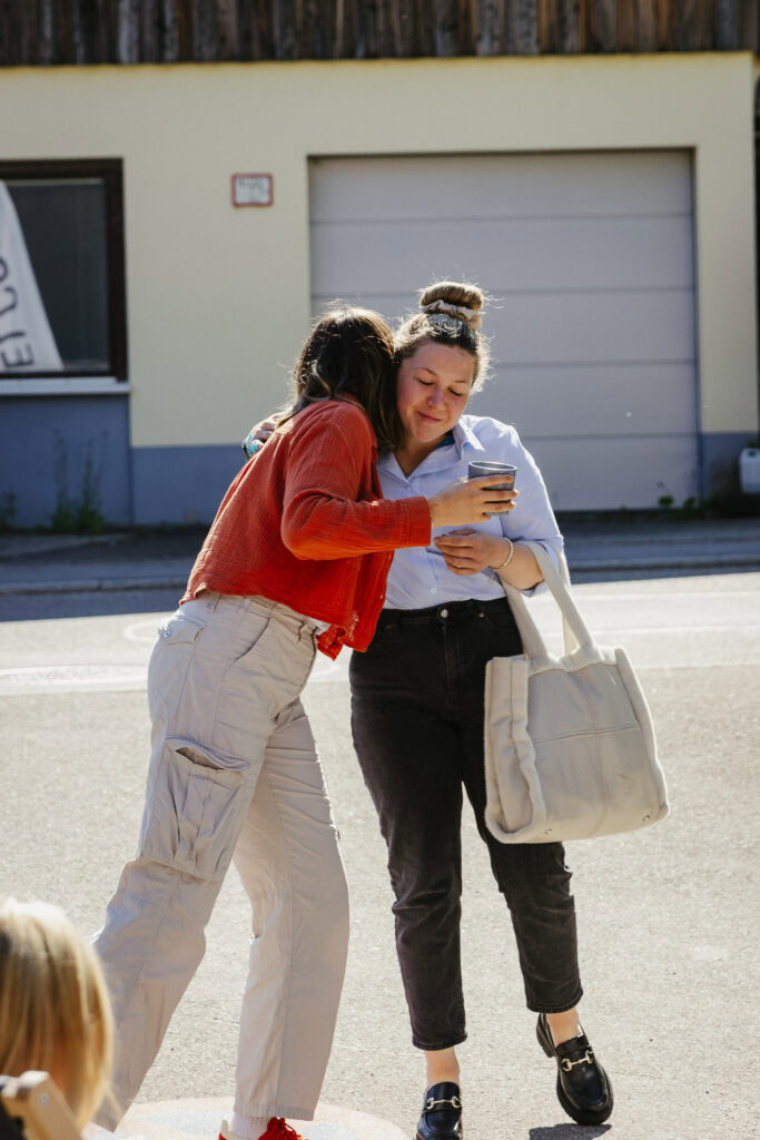 Zwei Frauen stehen auf der Straße, eine zeigt auf ihr Smartphone, die andere hält eine Tasche.