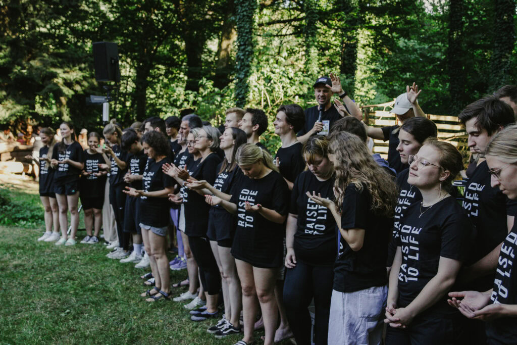 Menschen stehen in einer Reihe im Park, einige haben die Hände erhoben, andere halten Kerzen.