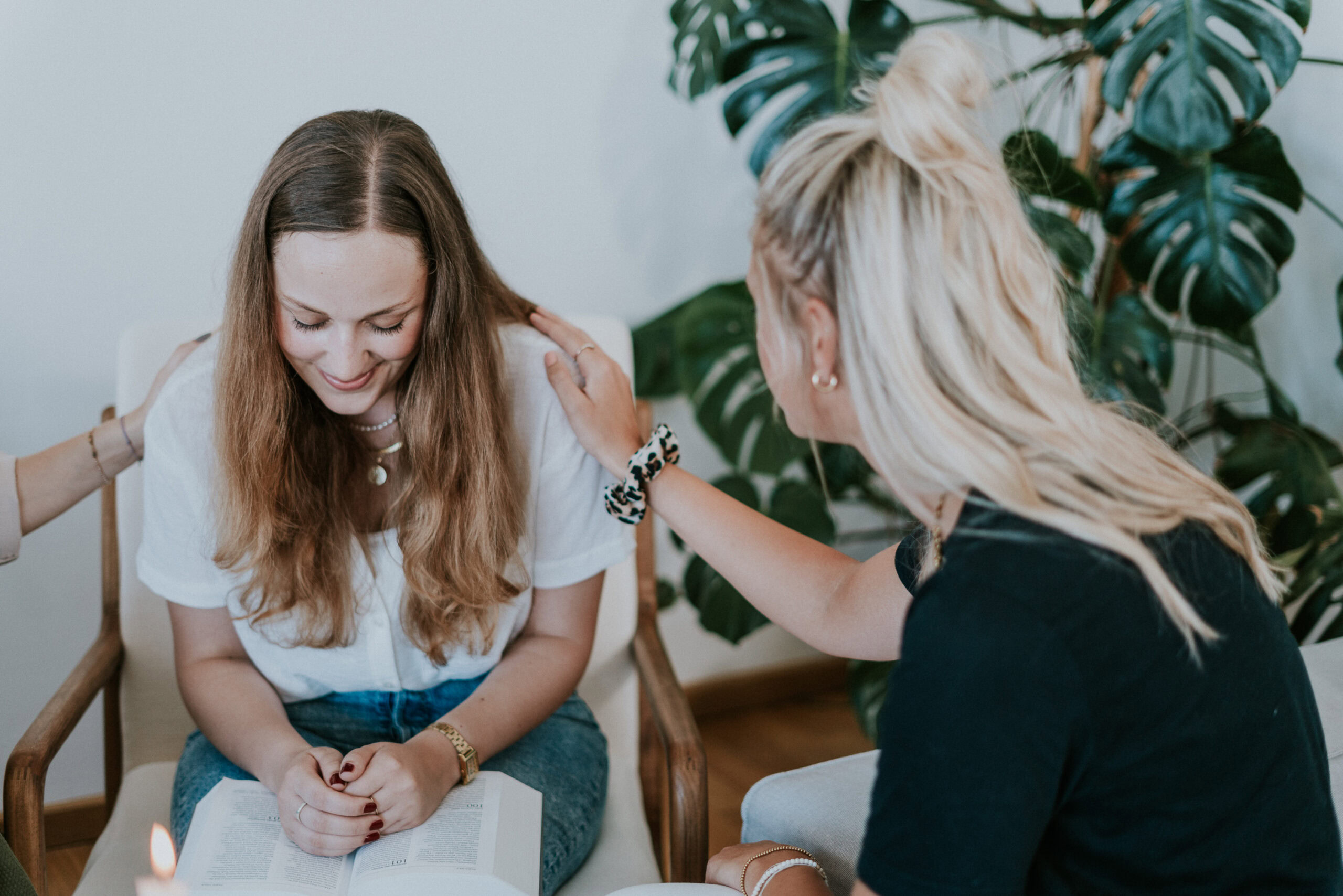 Zwei Frauen sitzen auf Stühlen, eine berührt die Schulter der anderen, während sie sprechen.