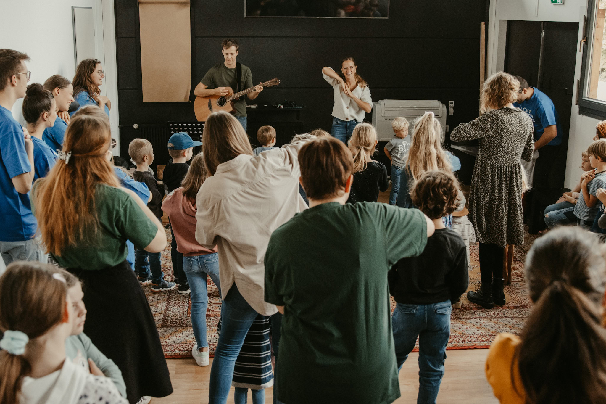 Zwei Musiker spielen Gitarre und Flöte vor einer Gruppe von Kindern und Erwachsenen in einem Raum.