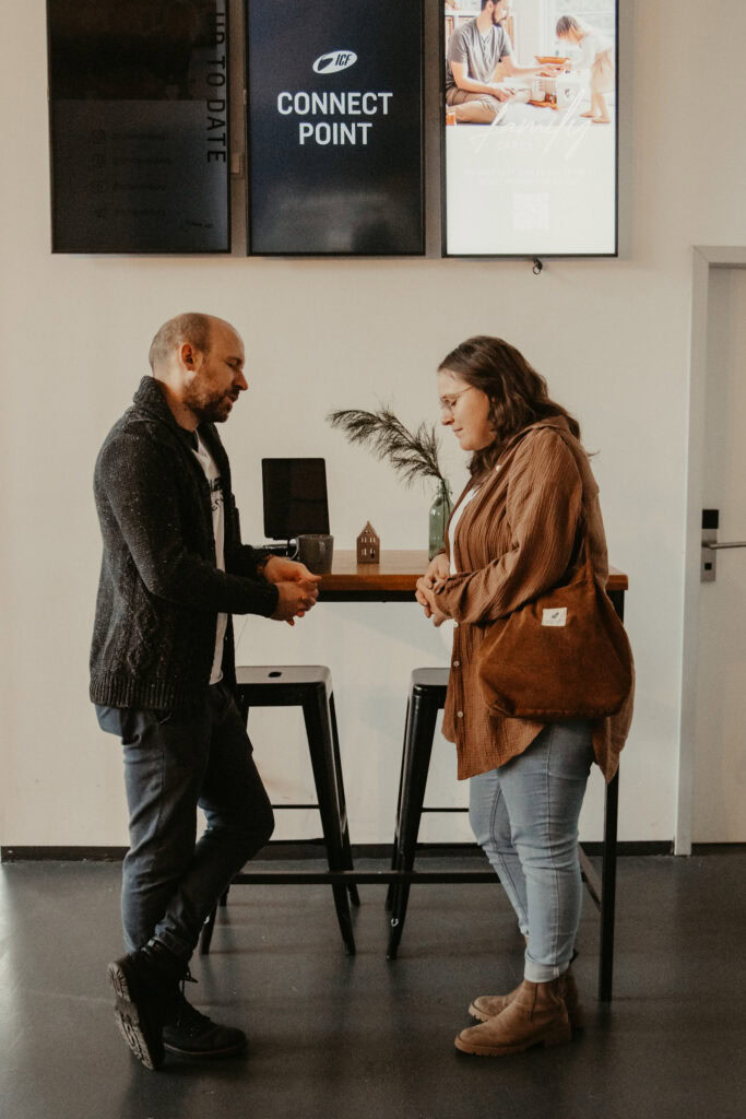 Zwei Menschen tauschen sich freundlich im Gemeindezentrum aus, im Hintergrund Bildschirme mit "Connect Point" und Gemeinschaftsaktivitäten.
