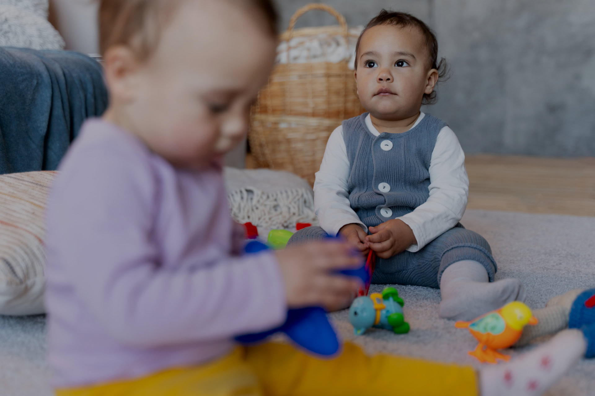 Zwei Kinder spielen auf einem Teppich mit buntem Spielzeug, eines schaut nach vorne.