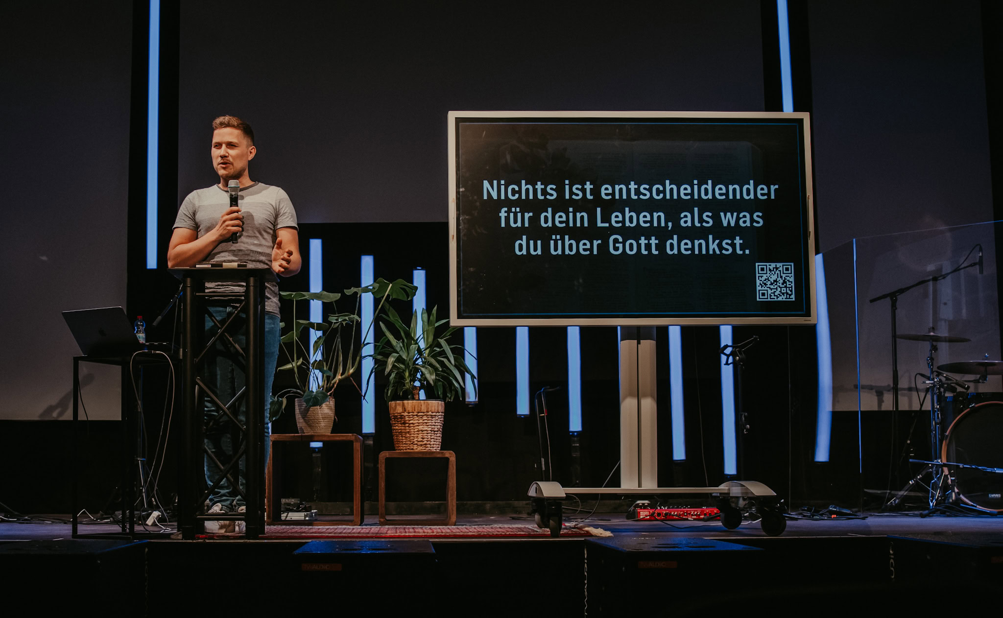 Mann steht an einem Pult auf einer Bühne, neben einem Bildschirm mit Text.