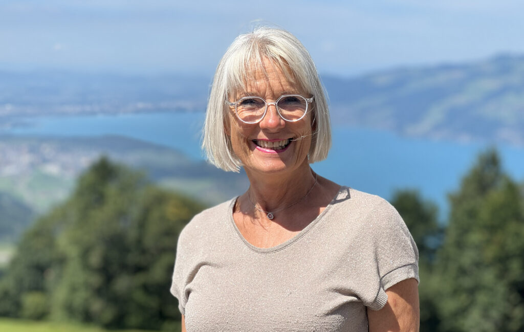 Eine ältere Frau mit weißen Haaren und Brille lächelt in die Kamera, im Hintergrund sind Bäume, Wass.