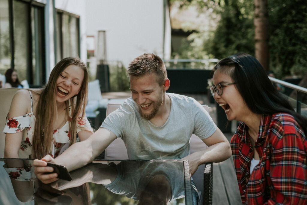 Drei Jugendliche sitzen an einem Tisch im Garten, lachen und schauen auf ein Smartphone.