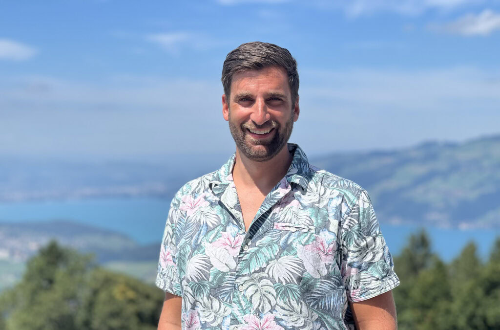Mann mit kurzen braunen Haaren und Bart, trägt ein buntes Blumen-Hemd, steht im Freien vor einer Lan.