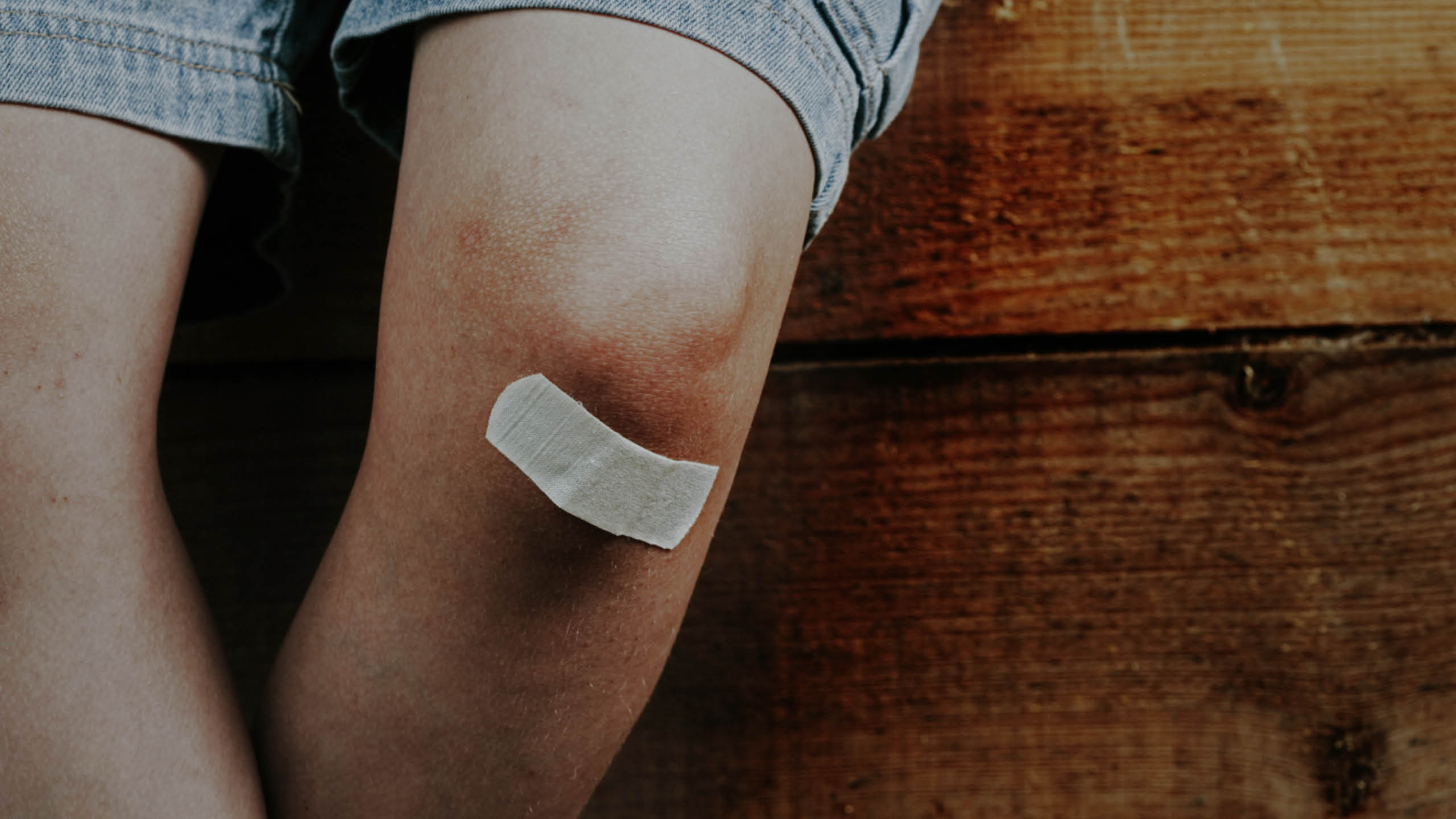 Nahaufnahme eines Knies mit Bluterguss und Verband, Holzoberfläche im Hintergrund.