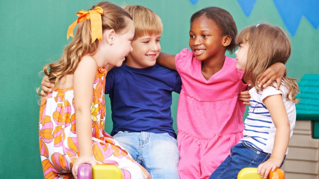 Vier lachende Kinder im Kreis, umarmen sich, im Hintergrund bunte Wand.
